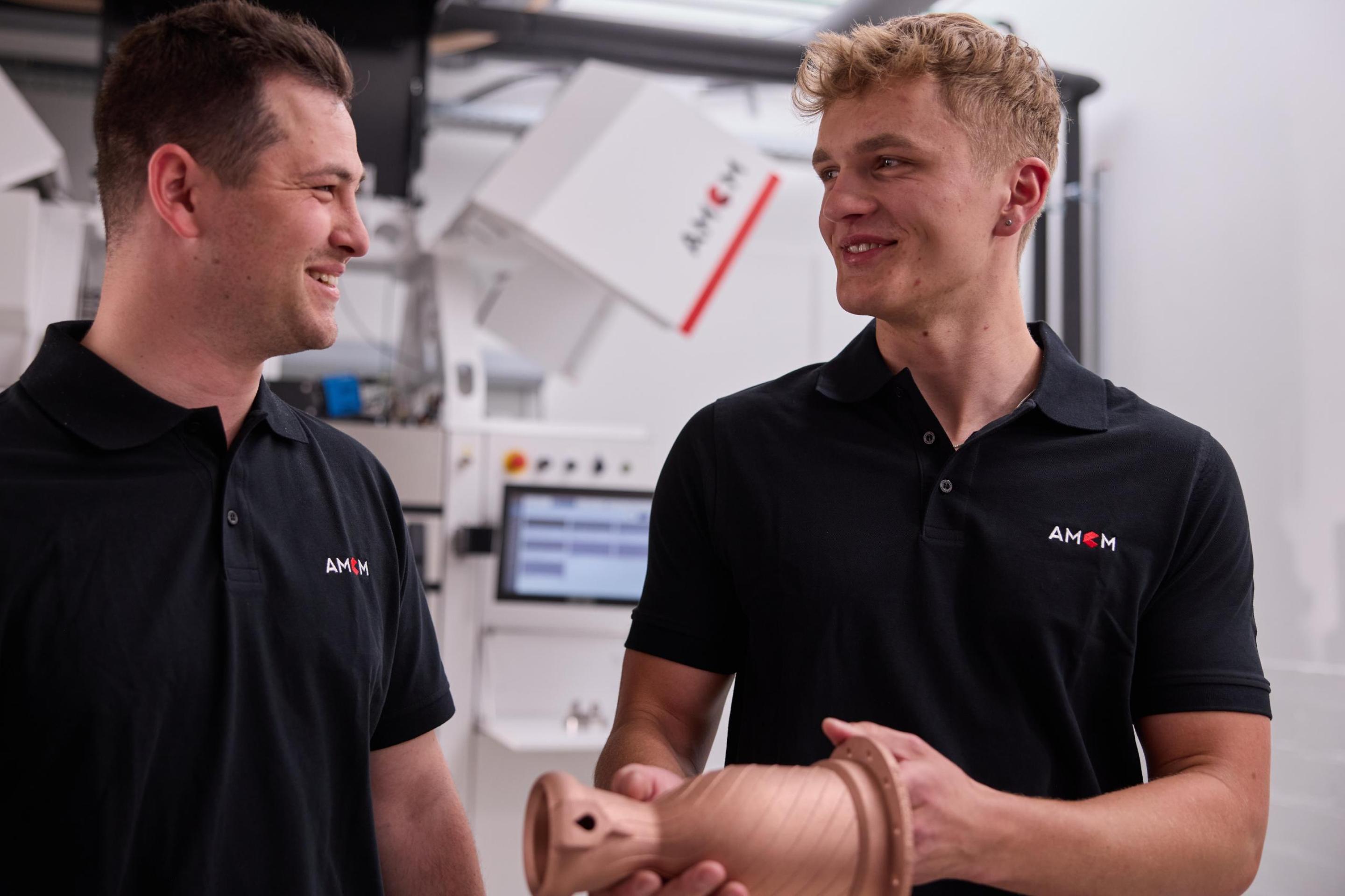 Two AMCM Employees with Copper Part in Front of AMCM Machine